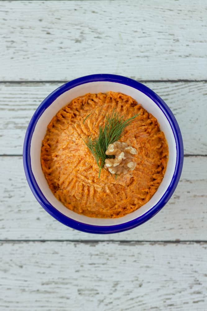 Orange vegan Turkish carrot dip inside blue lined, white enamel bowl with dill and walnut.